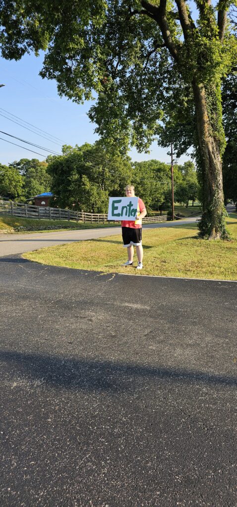 A student holding an “Enter” sign outdoors on a sunny driveway