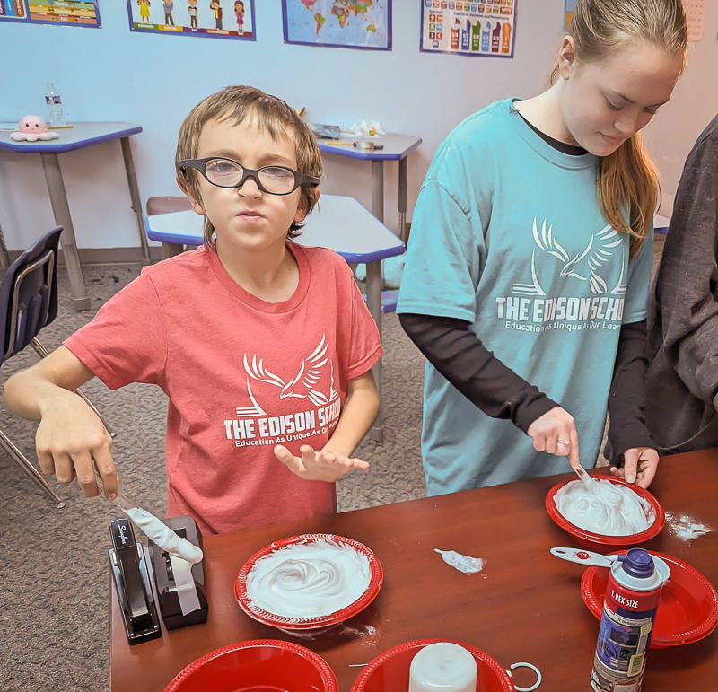 Two students in Edison School T-shirts spreading shaving cream on red plates during a class activity