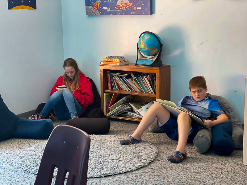 Two students reading on floor cushions beside the classroom bookshelf